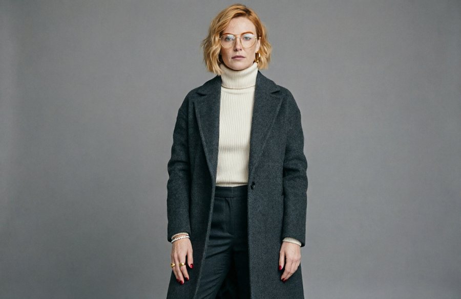 She wears a charcoal felted-wool longline coat with a high funnel collar standing clear of her jaw, one clean…